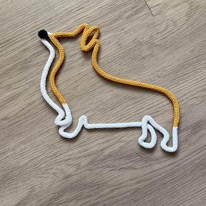 May include: A white and yellow knitted rope is shaped into the outline of a dog. The dog's nose is black.