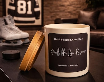 May include: A cream-colored candle with a wooden lid, featuring a black label with white text that reads "Smells like Ilya Rozanov." The candle is next to a hockey puck and ice skates. A framed hockey jersey is in the background.