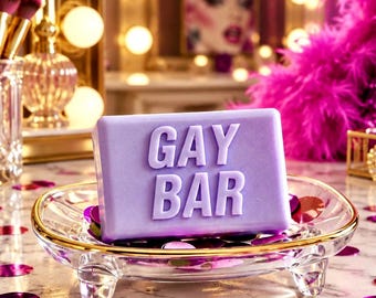 May include: A rectangular lavender-colored bar of soap with the words GAY BAR embossed in large capital letters on the front. The soap rests in a clear glass dish with a gold rim, surrounded by scattered purple and magenta circular confetti.