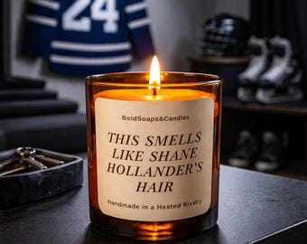 May include: A lit candle in a clear glass jar with a label that reads "THIS SMELLS LIKE SHANE HOLLANDER'S HAIR." The candle is handmade and part of a heated rivalry. A hockey jersey with the number 24 is in the background.