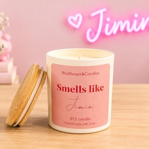 Può includere: Una candela bianca in un barattolo di vetro con un coperchio in legno chiaro. L'etichetta è rosa con testo rosso che recita BoldSoaps&Candles, Smells like Jimin, e BTS candle Handmade with love. La candela è posizionata su una superficie in legno.