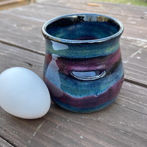 May include: A handmade ceramic tumbler with a vibrant, swirling glaze in shades of blue, green, and purple. The tumbler has a small handle and sits next to a white egg. The pottery is likely for drinking.