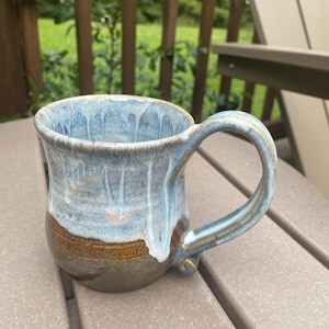 May include: A ceramic mug featuring a two-tone glaze with a dark brown base and a light blue upper section. The blue glaze drips down over the brown surface, creating a textured appearance. The mug includes a curved handle attached to the side.