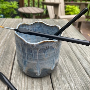 May include: A ceramic cup with a wavy rim and a blue and white drip glaze finish. Two black paintbrushes with orange tips are resting across the top of the cup, and a third matching paintbrush is positioned on the surface next to the cup.