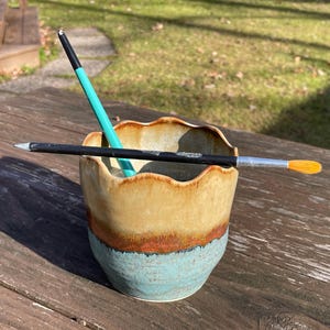 May include: A handmade ceramic cup with a wavy rim, featuring a color gradient of tan, orange, and turquoise. A teal pencil and a paintbrush with an orange tip rest inside the cup, and a black pencil rests on the rim.