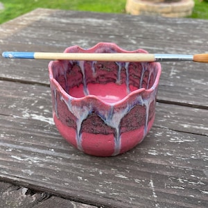 May include: A handmade ceramic bowl with a scalloped rim, featuring a vibrant pink and burgundy glaze with white and blue drips. A paintbrush with a wooden handle and orange bristles rests across the bowl.