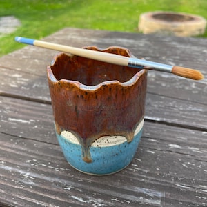 May include: A handmade ceramic cup with a brown and blue glaze, featuring a wavy rim and a paint brush resting on top. The brush has a wooden handle and a soft, orange-colored bristle head. The cup is approximately 4 inches tall.