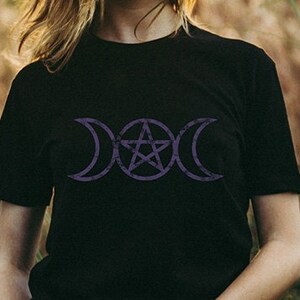 May include: A woman wearing a black t-shirt with a purple triple moon and pentagram design. She is standing in a field of tall grass.