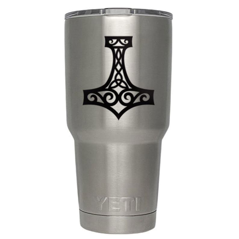 K&ouml;nnte beinhalten: Ein Edelstahlbecher mit geb&uuml;rsteter Metalloberfl&auml;che und transparentem Deckel. Auf der Vorderseite befindet sich eine schwarze Grafik eines Mjolnir-Hammers mit komplexen keltischen Knotenmustern. Der Markenname YETI ist am Boden eingepr&auml;gt.