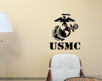 Könnte beinhalten: Ein schwarz-weißer Wandaufkleber mit dem Emblem des United States Marine Corps, einschließlich eines Adlers, einer Erdkugel und eines Ankers, mit dem Text "USMC" darunter. Der Aufkleber ist auf einer beigefarbenen Wand mit einem weißen Stuhl und einer Lampe im Hintergrund angebracht.