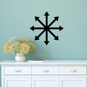 May include: A black eight-pointed star with arrows pointing outwards, centered on a light blue wall. A white dresser with two drawers and two doors is in the foreground. A vase with white and green flowers sits on top of the dresser.