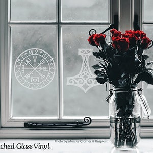 K&ouml;nnte beinhalten: Ein klares Glasgef&auml;&szlig; mit dunkelroten Rosen steht vor einer Fensterscheibe mit wei&szlig;en Vinyl-Aufklebern in &Auml;tzglasoptik. Die Motive auf der Glasoberfl&auml;che zeigen einen kreisf&ouml;rmigen Vegvisir-Kompass, einen Mj&ouml;lnir-Hammer und ein Valknut-Symbol.