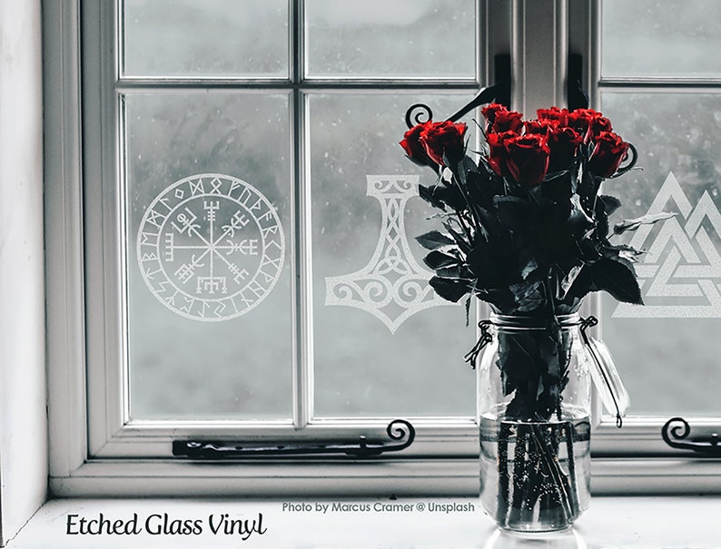 K&ouml;nnte beinhalten: Ein klares Glasgef&auml;&szlig; mit dunkelroten Rosen steht vor einer Fensterscheibe mit wei&szlig;en Vinyl-Aufklebern in &Auml;tzglasoptik. Die Motive auf der Glasoberfl&auml;che zeigen einen kreisf&ouml;rmigen Vegvisir-Kompass, einen Mj&ouml;lnir-Hammer und ein Valknut-Symbol.