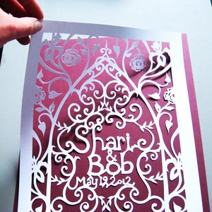 May include: White paper cut-out design with a burgundy background. The design features a floral pattern with the names "Shari & Bob" and the date "May 19, 2012".