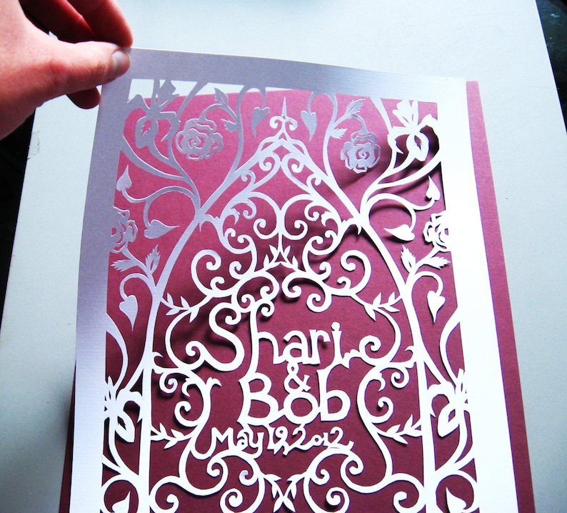 May include: White paper cut-out design with a burgundy background. The design features a floral pattern with the names "Shari & Bob" and the date "May 19, 2012".