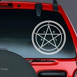 May include: White vinyl decal featuring a pentagram within a double circle, with Hebrew letters around the perimeter and within the star. The decal is affixed to the rear window of a red vehicle. The text "Arwen Designs" is visible in the lower left corner.