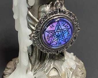 May include: A silver necklace with a pentagram pendant. The pendant is a glass cabochon with a purple and blue gradient background and a black pentagram with the words "TRA" "GRA" "TE" "MA" "TON" around it.