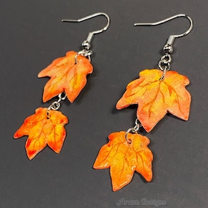 May include: A pair of dangle earrings featuring orange and yellow maple leaf charms. The leaves have a glossy finish and are suspended from silver-colored fish hook ear wires. The earrings are a fall-themed accessory.