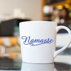 Puede incluir: Taza de cer&aacute;mica blanca con una inscripci&oacute;n azul en cursiva que dice "Namaste".