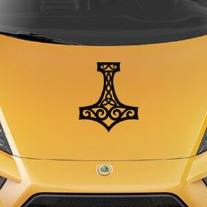K&ouml;nnte beinhalten: Ein schwarzer Vinyl-Aufkleber mit einem stilisierten Mjolnir-Hammer-Design und komplexen keltischen Knotenmustern, der mittig auf einer leuchtend gelben Motorhaube angebracht ist. Die Grafik ist einfarbig schwarz und zeigt symmetrische Schn&ouml;rkel.