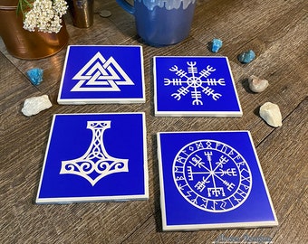 May include: Set of four blue ceramic coasters with white Norse symbols. The coasters feature a Vegvisir, a Valknut, Mjolnir, and a compass rose.