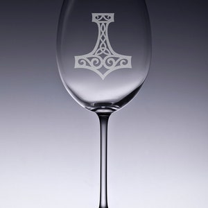 K&ouml;nnte beinhalten: Ein klares Weinglas aus Glas mit einem mattierten, ge&auml;tzten Design eines Mjolnir-Hammers auf dem Kelch. Der Stiel ist schlank und mit einem runden Fu&szlig; verbunden. Das Glas ist transparent mit einer glatten Oberfl&auml;che und einem Knotenmuster.