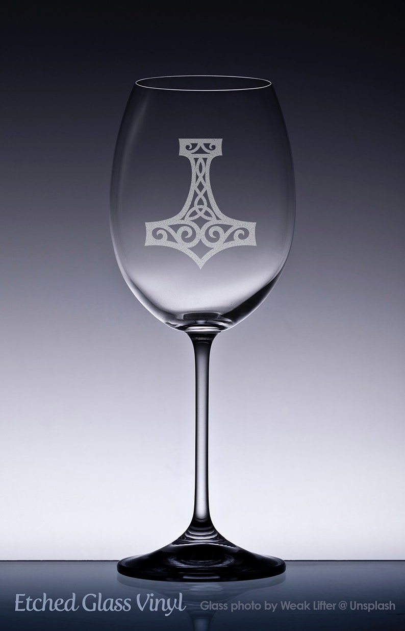 K&ouml;nnte beinhalten: Ein klares Weinglas aus Glas mit einem mattierten, ge&auml;tzten Design eines Mjolnir-Hammers auf dem Kelch. Der Stiel ist schlank und mit einem runden Fu&szlig; verbunden. Das Glas ist transparent mit einer glatten Oberfl&auml;che und einem Knotenmuster.