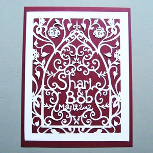 May include: White paper cut-out design on a burgundy background. The design features a floral arch with the names "Shari & Bob" and the date "May 19, 2012".