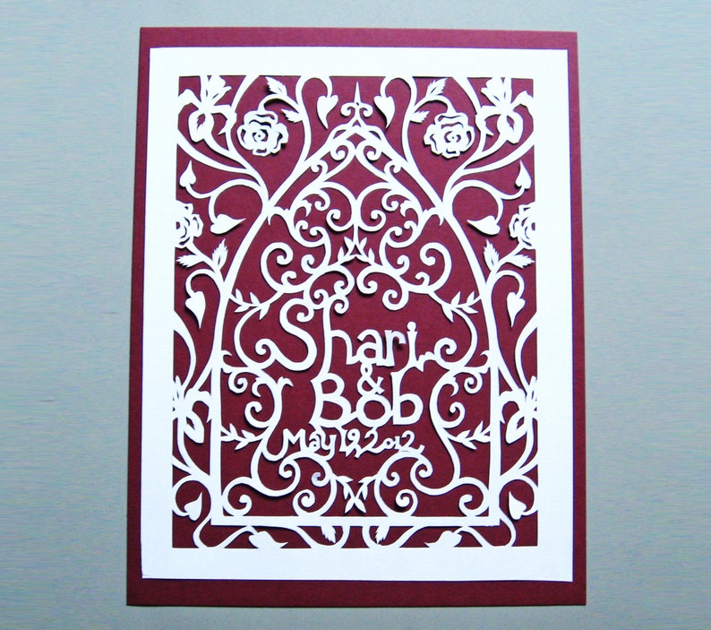 May include: White paper cut-out design on a burgundy background. The design features a floral arch with the names "Shari & Bob" and the date "May 19, 2012".