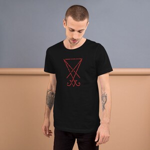 Pode incluir: Uma camiseta preta com um s&iacute;mbolo vermelho de pentagrama com uma cruz dentro. O s&iacute;mbolo &eacute; um s&iacute;mbolo comum associado ao satanismo.