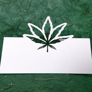 May include: White card with a cut-out marijuana leaf design. The card is on a green background.