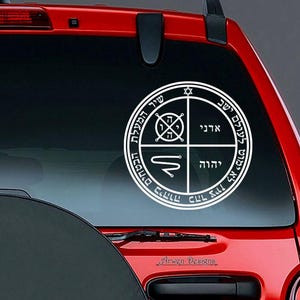 May include: A white circular decal with Hebrew text and symbols, affixed to a red vehicle window. The design features a central star and various inscriptions within concentric circles and quadrants. The decal is a product of Arwen Designs.