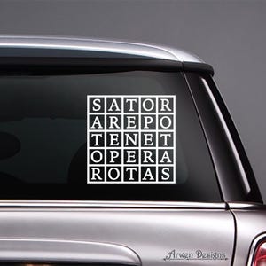 May include: A white vinyl car decal featuring the text "SATOR AREPO TENET OPERA ROTAS" arranged in a square grid. The decal is affixed to the rear window of a silver car. The words are in a serif font.