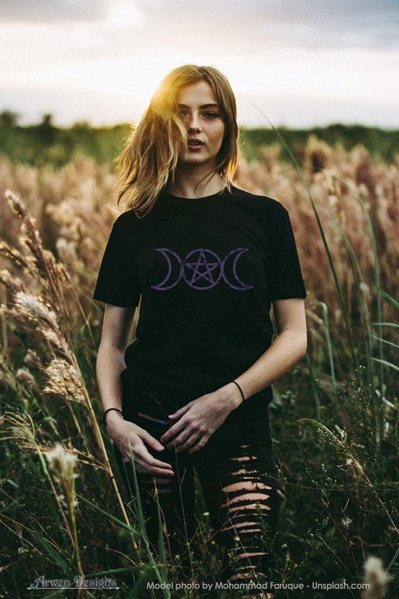 May include: A woman wearing a black t-shirt with a purple triple moon and pentagram design. She is standing in a field of tall grass.