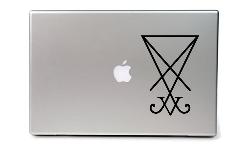 May include: A silver laptop with a black sticker of the sigil of Satan on the lid. The sigil is a pentagram with a cross inside and the letters 'XV' below it.