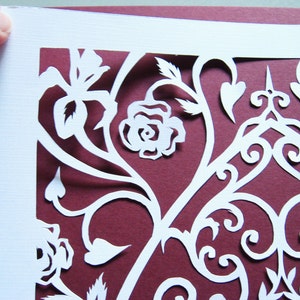 May include: White paper cut-out design with intricate floral patterns and a rose on a burgundy background.