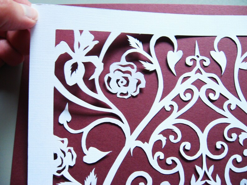 May include: White paper cut-out design with intricate floral patterns and a rose on a burgundy background.