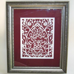 May include: A white paper cut-out design framed in a silver frame. The design features a floral pattern with the names "Shari & Bob" and the date "May 19, 2012".