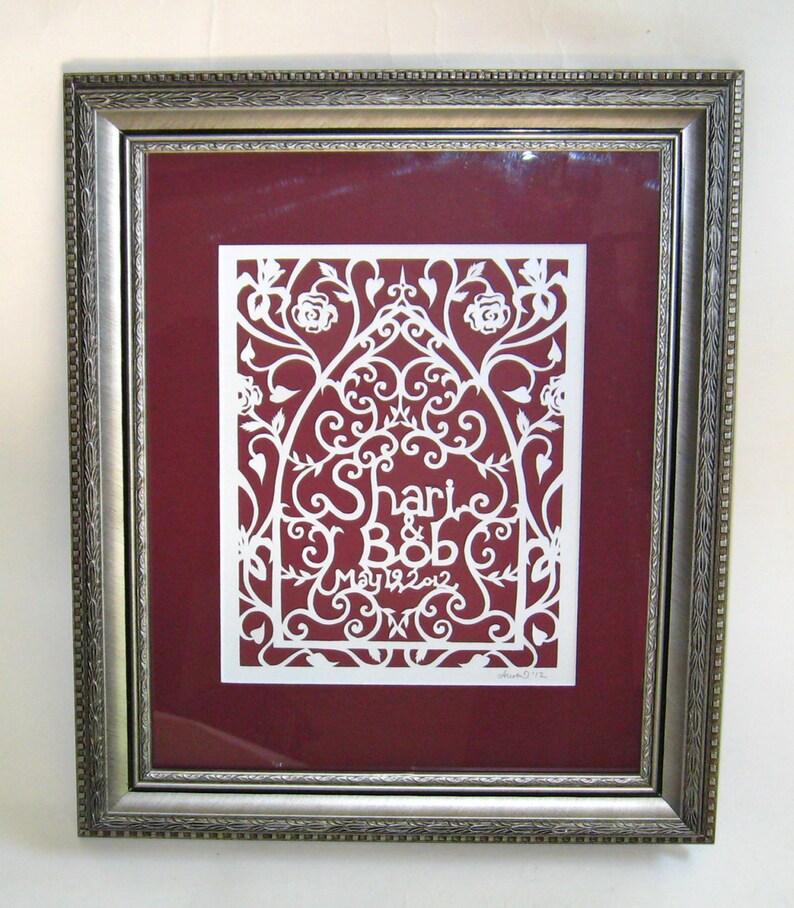 May include: A white paper cut-out design framed in a silver frame. The design features a floral pattern with the names "Shari & Bob" and the date "May 19, 2012".