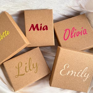 Op de afbeelding: Vijf kleine, vierkante kraftpapieren dozen met namen erop gedrukt in verschillende kleuren. De namen zijn Charlotte, Mia, Olivia, Lily en Emily. De dozen zijn gerangschikt op een wit, getextureerd oppervlak.