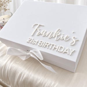 Könnte beinhalten: Eine rechteckige weiße Geschenkbox mit einer weißen Schleife auf der Vorderseite. Der Deckel zeigt erhabene weiße Schriftzüge mit dem Text Frankie's 21st BIRTHDAY in einer Kombination aus Schreib- und Blockschrift für ein klares Design.