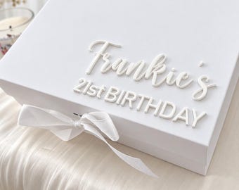 Puede incluir: Una caja de regalo rectangular blanca con un lazo blanco atado en la parte delantera. La tapa muestra letras blancas en relieve que dicen Frankie's 21st BIRTHDAY en una combinación de fuentes cursivas y de bloque, creando un aspecto limpio.