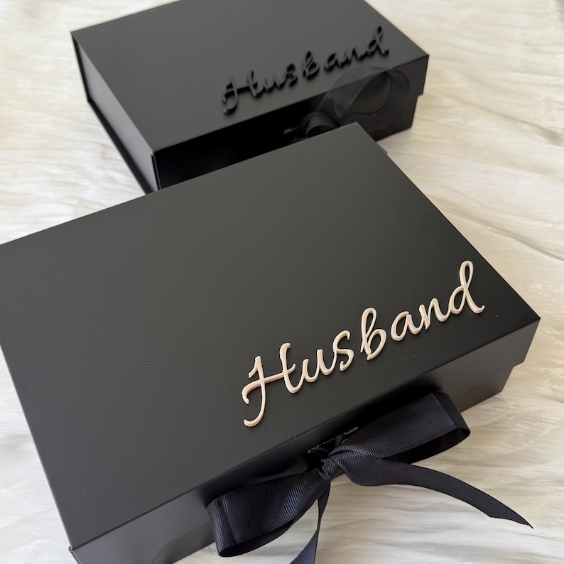 May include: Two rectangular black gift boxes with hinged lids. Each box features the word Husband in cursive script on the lid. The front of each box is adorned with a black ribbon tied in a bow. One box has black lettering, while the other has light beige lettering.