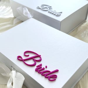 Puede incluir: Dos cajas de regalo rectangulares blancas con cierres magn&eacute;ticos y lazos de cinta blanca. La caja frontal presenta la palabra Bride en letras rosas fucsia texturizadas en relieve, mientras que la caja trasera muestra la palabra Bride en plata.