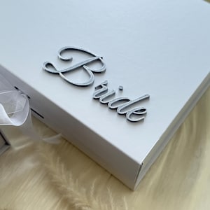 Puede incluir: Una caja de regalo rectangular blanca con la palabra Bride en una fuente cursiva plateada met&aacute;lica en relieve sobre la tapa. La caja incluye un cierre de cinta blanca en el lateral, ofreciendo una est&eacute;tica limpia para un contenedor de presentaci&oacute;n.