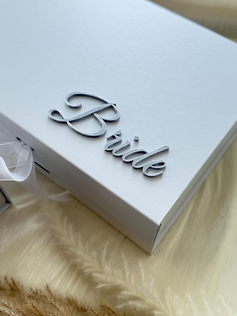 Puede incluir: Una caja de regalo rectangular blanca con la palabra Bride en una fuente cursiva plateada met&aacute;lica en relieve sobre la tapa. La caja incluye un cierre de cinta blanca en el lateral, ofreciendo una est&eacute;tica limpia para un contenedor de presentaci&oacute;n.