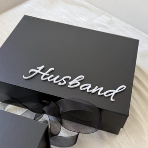 May include: A rectangular black gift box featuring the word Husband in white script lettering attached to the lid. The box is adorned with a black sheer ribbon tied in a bow at the front, and a portion of a second smaller black box is visible below.