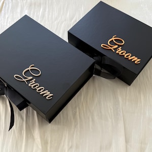 May include: Two rectangular black gift boxes with magnetic closures and black ribbon ties. Each box features the word Groom in a metallic gold script font attached to the lid. The boxes have a smooth matte finish and a structured design.