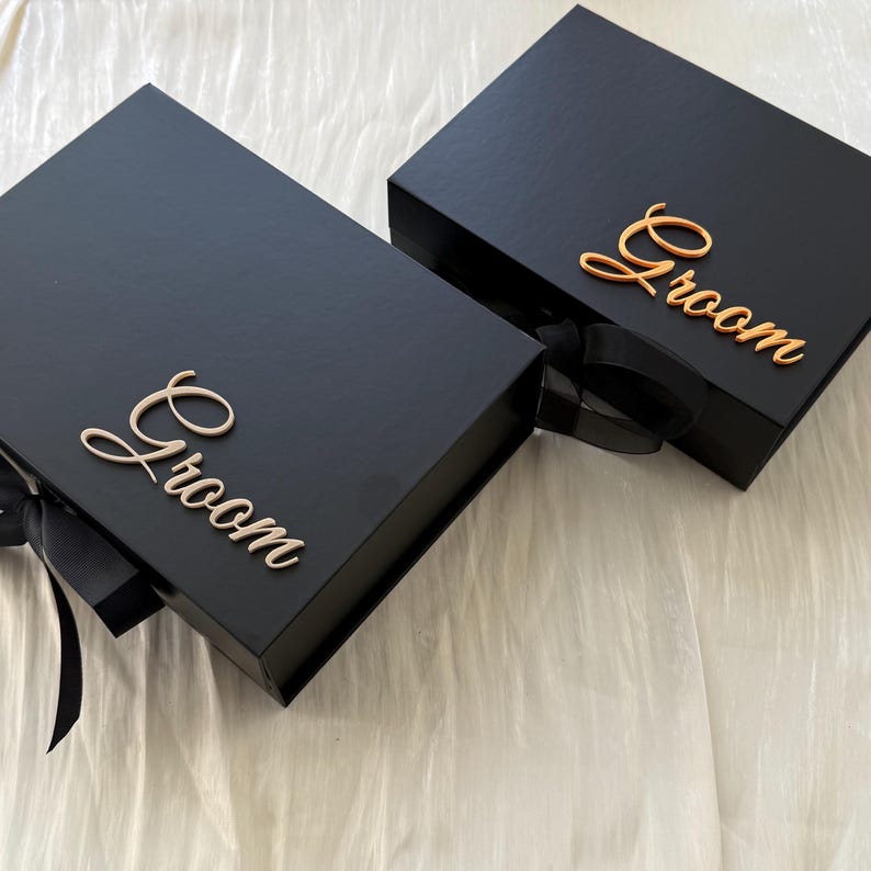 May include: Two rectangular black gift boxes with magnetic closures and black ribbon ties. Each box features the word Groom in a metallic gold script font attached to the lid. The boxes have a smooth matte finish and a structured design.