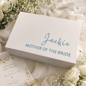 May include: A rectangular white gift box featuring the name Jackie in a light blue script font and the text MOTHER OF THE BRIDE in a light blue sans-serif font on the lid. The box has a smooth finish and a clean, minimalist design.